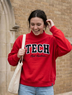 Student in the red Texas Tech Dark Horse “Campus Club” mock neck sweatshirt with black chenille TECH lettering, walking past a campus building with a canvas tote