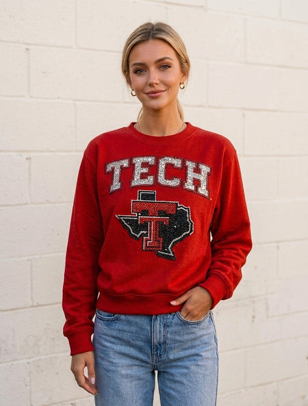 Woman wearing a red crewneck sweatshirt with TECH and the Lone Star Pride Logo in rhinestones, standing in front of a white cinderblock wall